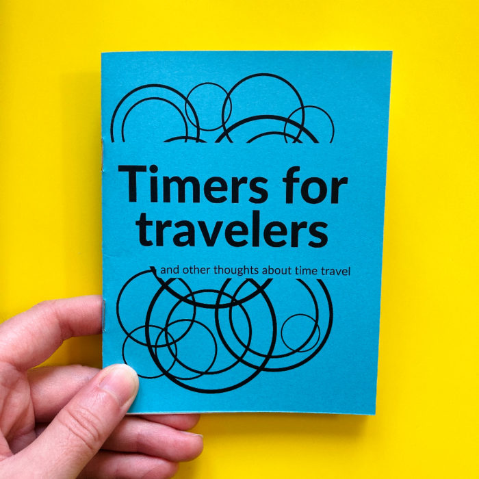 A hand holding "Timers for travelers" zine. The zine cover is blue paper. The title is printed in black. A pattern of overlapping circles is behind the title.