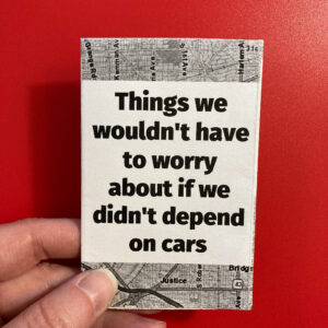 A hand holds a mini zine called "Things we wouldn't have to worry about if we didn't depend on cars." The text is printed in black on a white background. Above and below the text, there's a black and white map of Chicago streets.