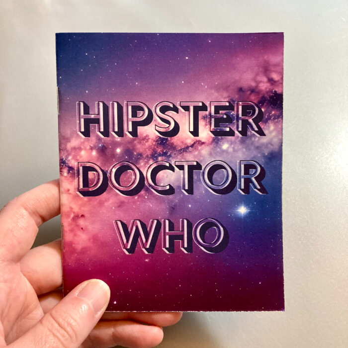 A hand holding the zine "Hipster Doctor Who." The zine cover has a purple, blue, and red galaxy image in the background. Text on top of the image says "Hipster Doctor Who" in all uppercase letters.