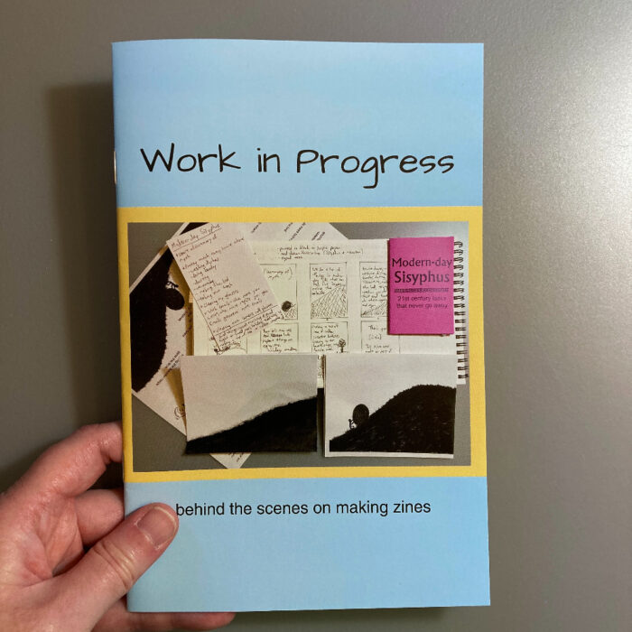 A hand holding the zine "Work in Progress." The cover of the zine has a light blue background. A photo on the cover shows an index card with notes, some illustrations of a man pushing a boulder up a hill, a notebook, and a purple zine titled "Modern-day Sisyphus." Below the photo, text says "behind the scenes on making zines."