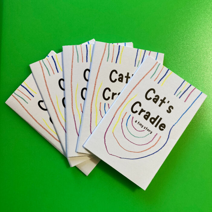 Five copies of the zine "Cat's Cradle: A tiny story," fanned out on a green background.