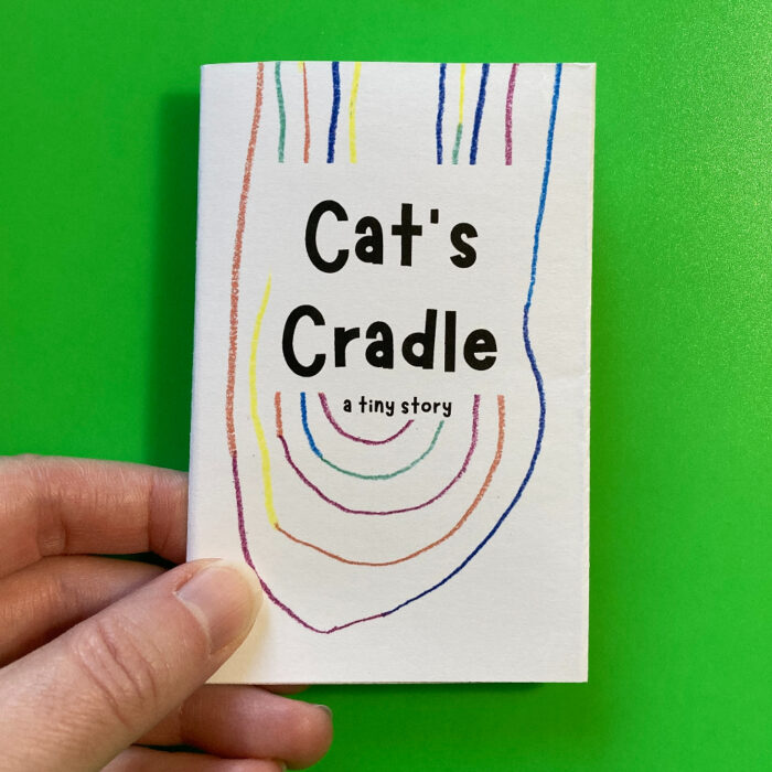 A hand holding the zine "Cat's Cradle: A tiny story." The cover includes the title of the zine. Behind the text, there are multicolored curved lines drawn with colored pencils.